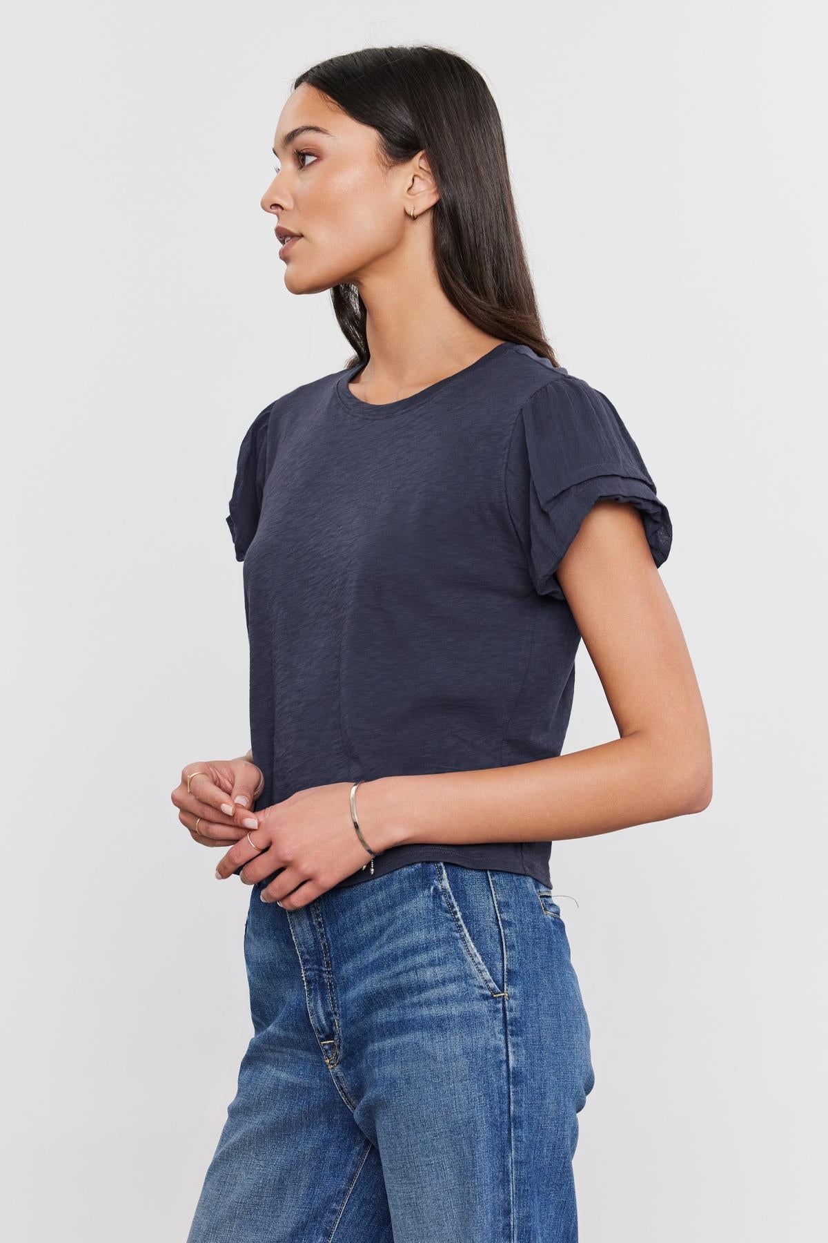   Profile view of a woman in a dark blue Della Tee by Velvet by Graham & Spencer with pleated sleeve detail and blue jeans, standing against a white background. 