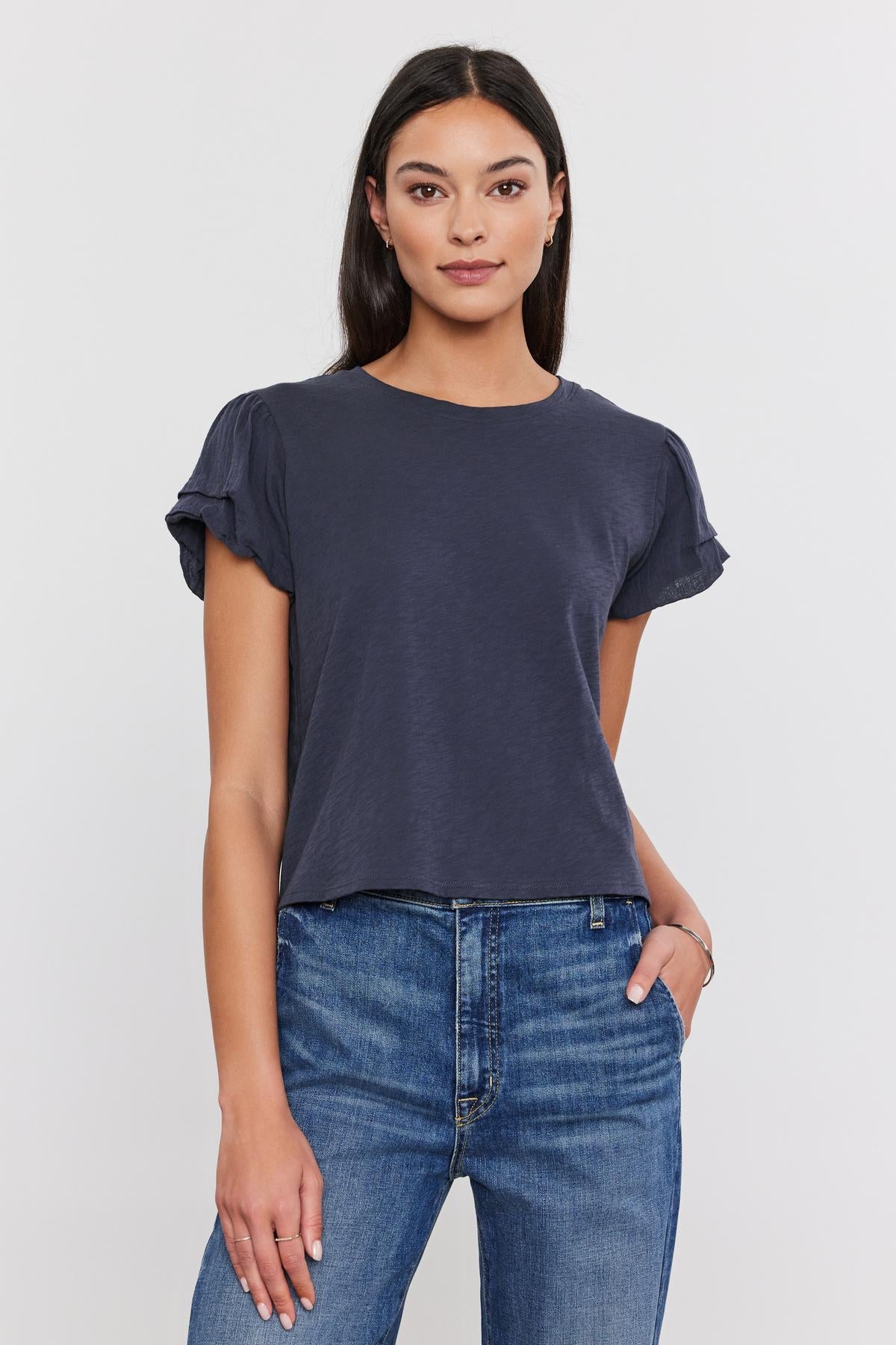 A woman in a Velvet by Graham & Spencer DELLA TEE with pleated sleeve detail and blue jeans standing against a white background, looking directly at the camera.-36805187272897