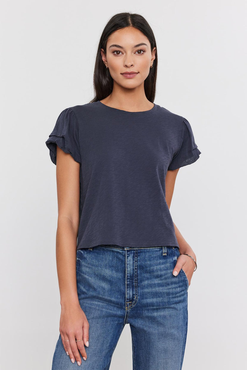 A woman in a Velvet by Graham & Spencer DELLA TEE with pleated sleeve detail and blue jeans standing against a white background, looking directly at the camera.