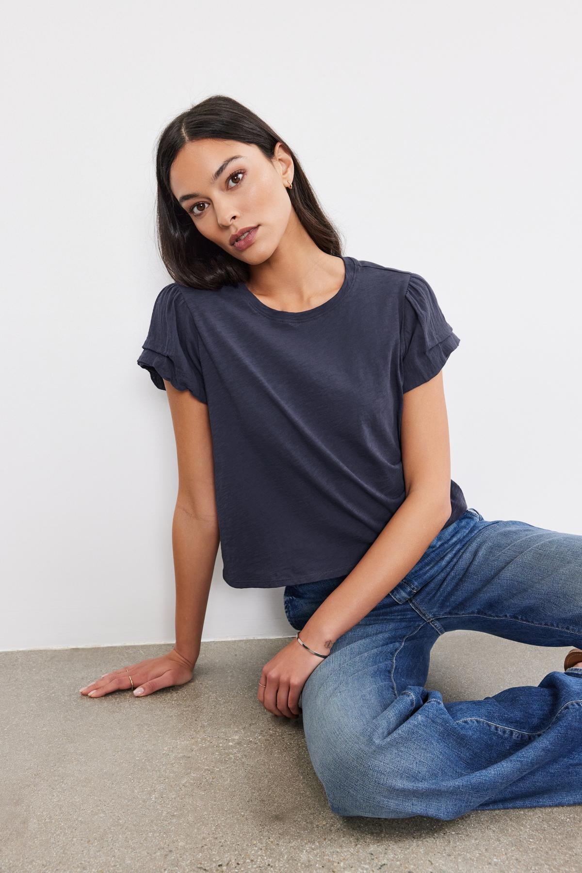   A woman in a grey Velvet by Graham & Spencer DELLA TEE with pleated sleeve detail and blue jeans sitting on the floor against a white background, looking at the camera. 