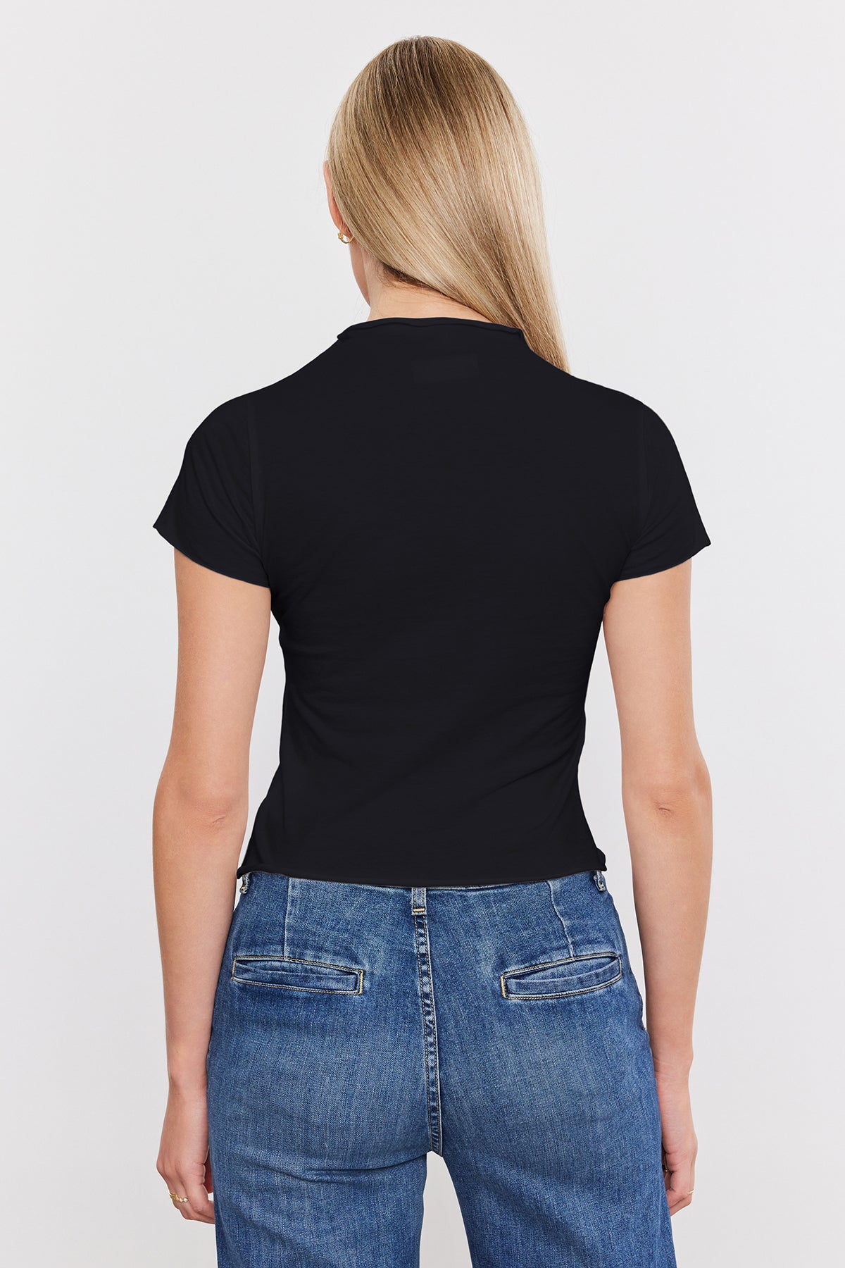   Woman viewed from behind wearing a black Velvet by Graham & Spencer JACKIE MOCK NECK TEE and blue jeans, standing against a white background. 
