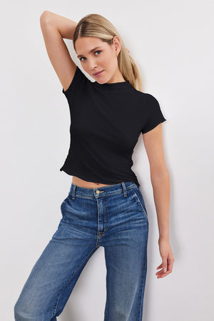 A woman posing with one hand on her head, wearing a black Velvet by Graham & Spencer JACKIE MOCK NECK TEE and blue jeans, against a plain white background.