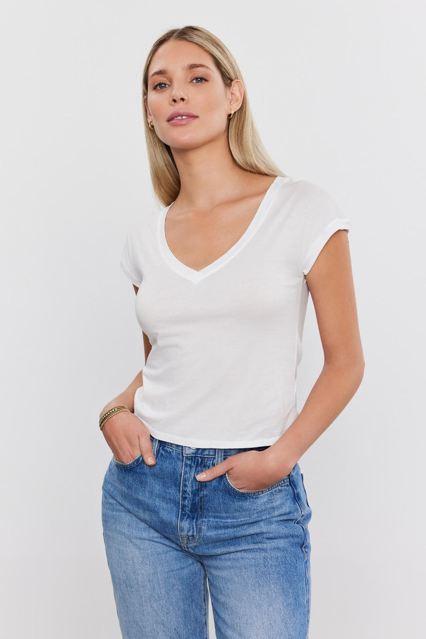 A woman wearing a white Velvet by Graham & Spencer TOBY TEE and blue jeans against a plain white background. she is posing with one hand in her pocket and looking at the camera.