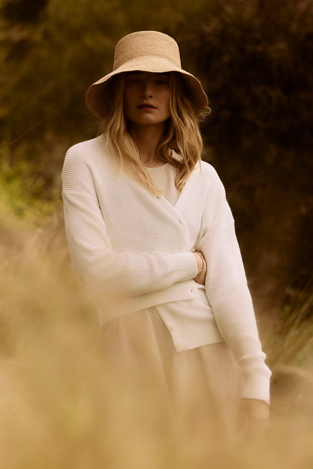   A woman in a white Velvet by Graham & Spencer TAVA cardigan and straw hat stands in a sunlit field, partially obscured by foreground foliage. 
