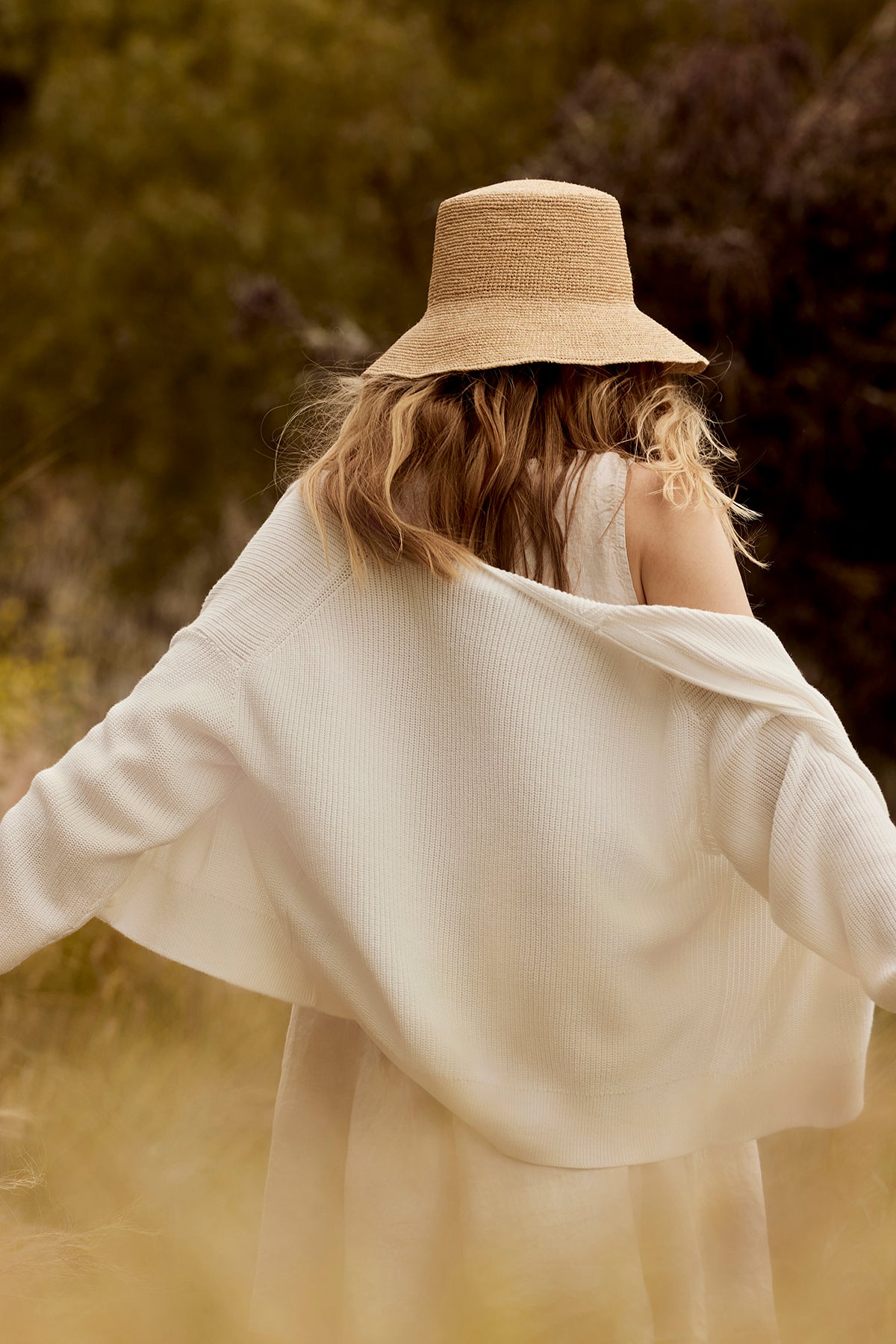 A woman with curly hair, seen from behind, wears a straw hat and a white flowing blouse in a sunlit, grassy field. She has draped over her shoulders a Velvet by Graham & Spencer Tava Cardigan.-36909783056577