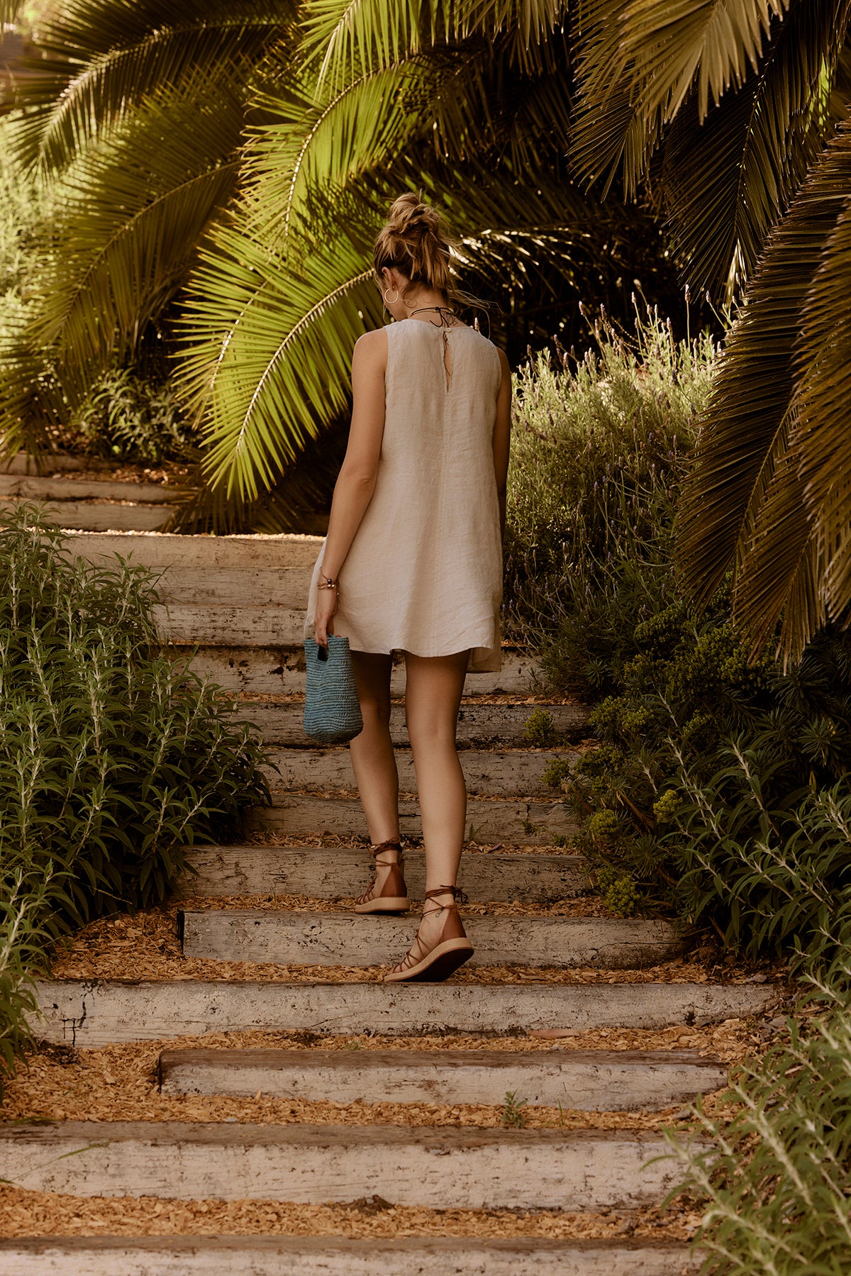 A woman in a Velvet by Graham & Spencer Tiffany Linen Dress walking up a stone staircase, surrounded by lush greenery, holding a blue bag.-36909787316417