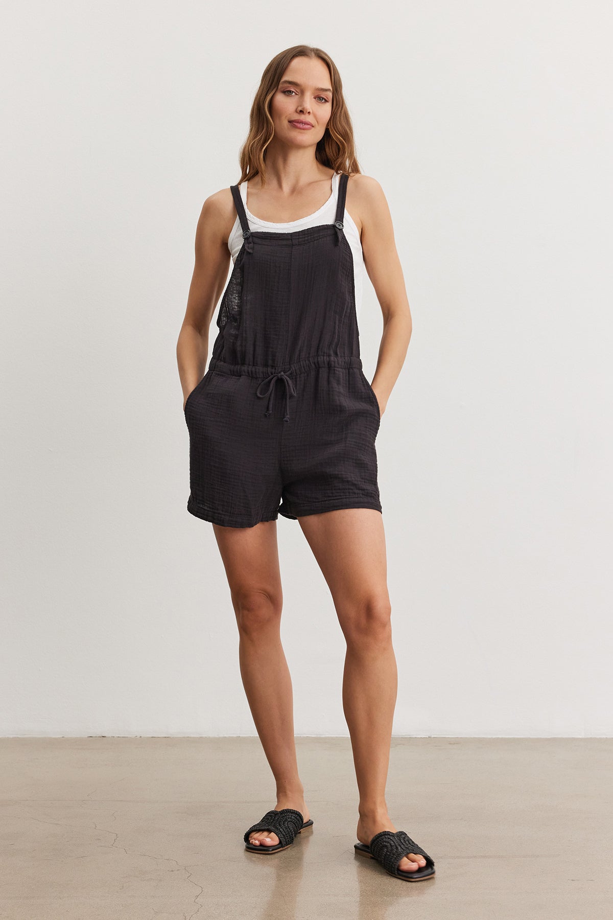 A woman stands in a studio, wearing a black CYNDEE COTTON GAUZE ROMPER by Velvet by Graham & Spencer with drawstring waist and black slide sandals, smiling subtly.-36909553451201