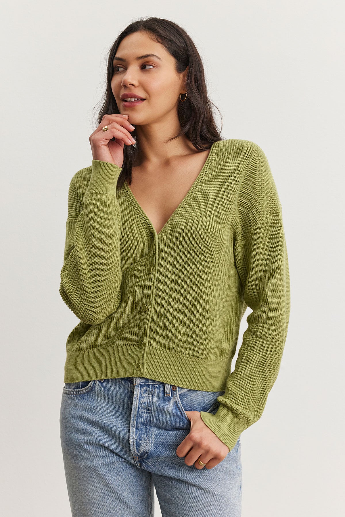   Woman in a green Velvet by Graham & Spencer Tava cardigan and blue jeans standing against a white background, touching her chin thoughtfully. 