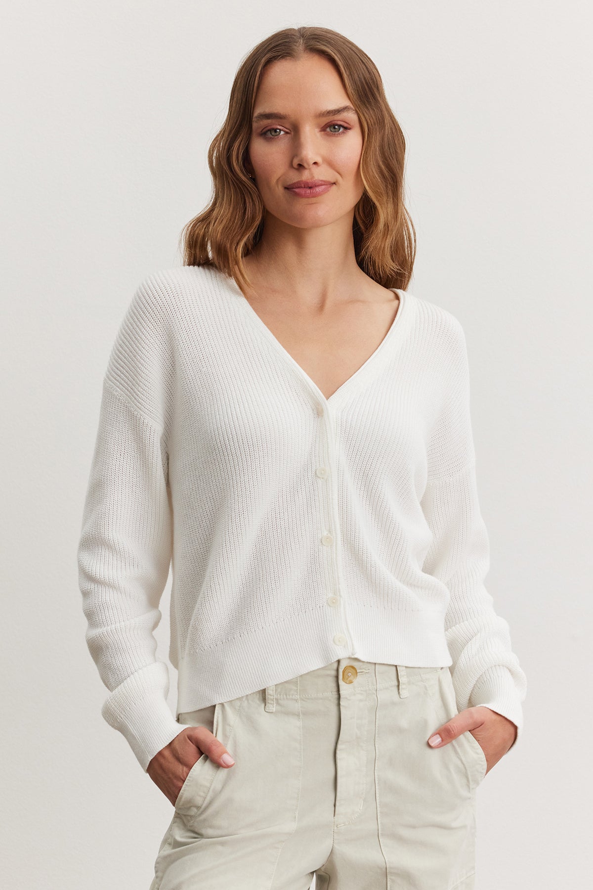 A woman with shoulder-length brown hair wearing a white TAVA Cardigan by Velvet by Graham & Spencer and beige trousers, standing against a plain background.-36909564264641