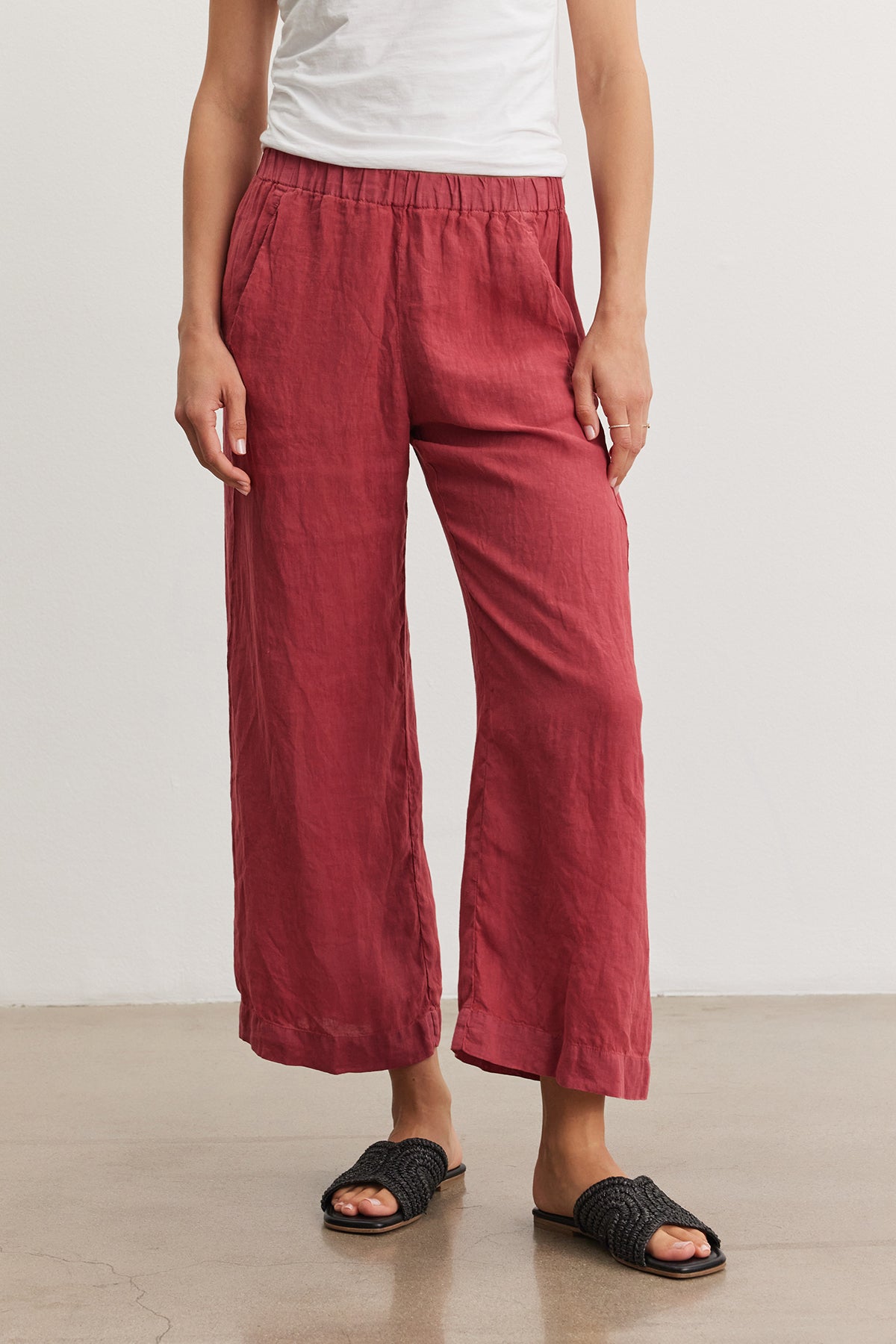 A person wearing the LOLA LINEN PANT by Velvet by Graham & Spencer with an elastic waist and a white top paired with black slide sandals, shown from the waist down in a neutral setting.-36909573308609