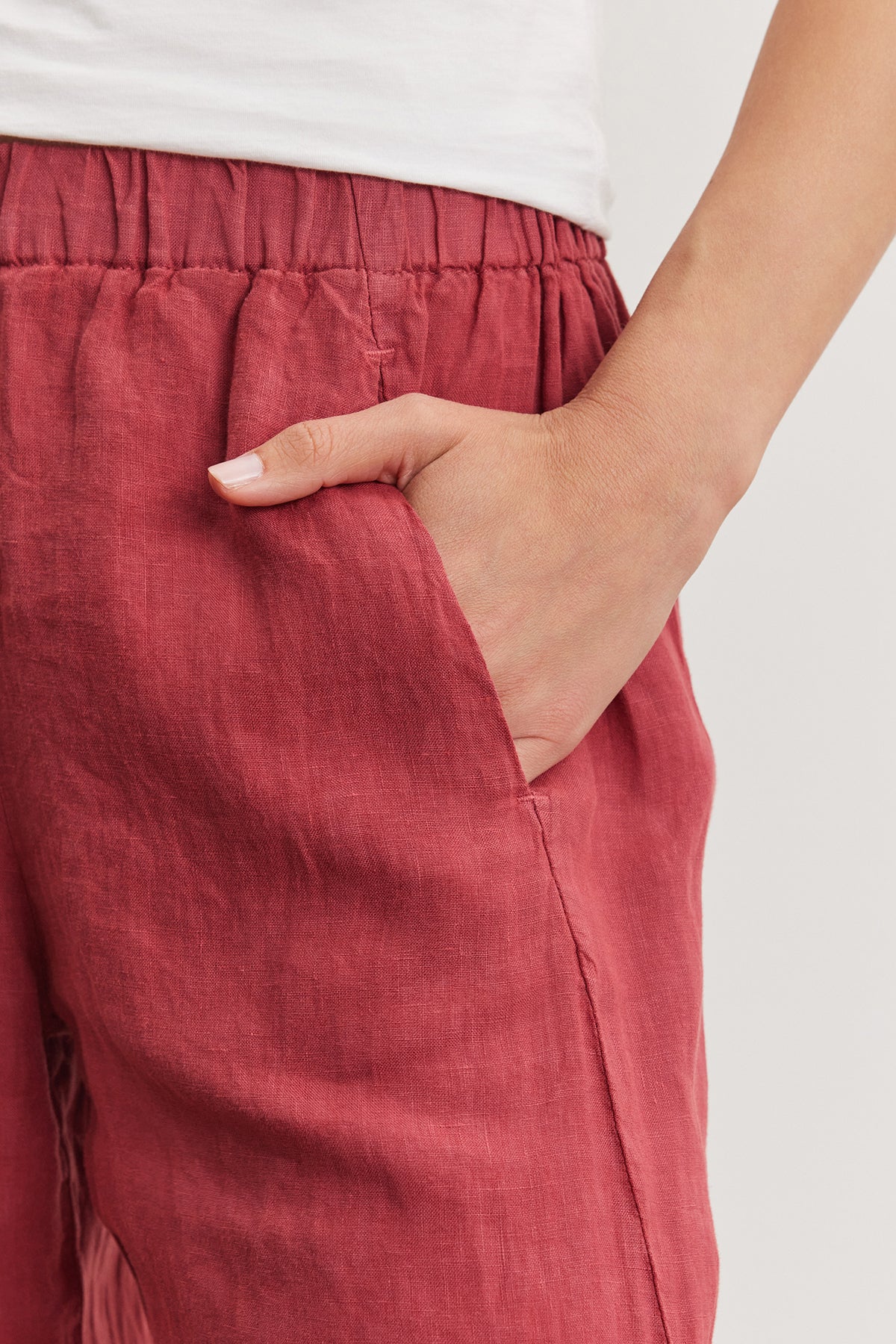 Close-up of a person's hand with a white manicure partially inserted into the pocket of an ankle crop red Velvet by Graham & Spencer Lola Linen Pant.-36909573406913
