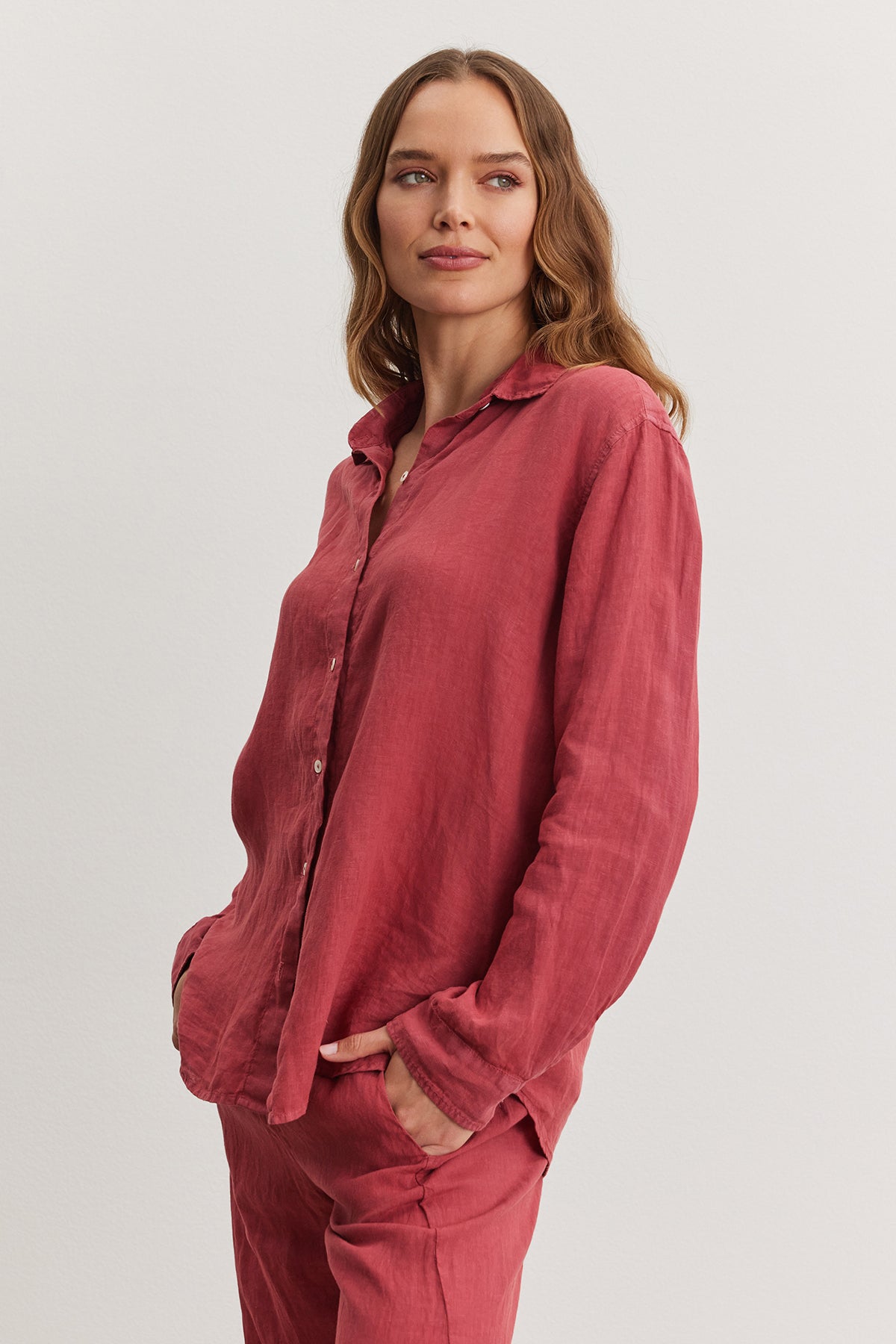 Woman in a relaxed fit red Velvet by Graham & Spencer Willow Linen Button-Up Shirt and pants, standing with one hand in pocket, looking at the camera with a slight smile.-36909598310593