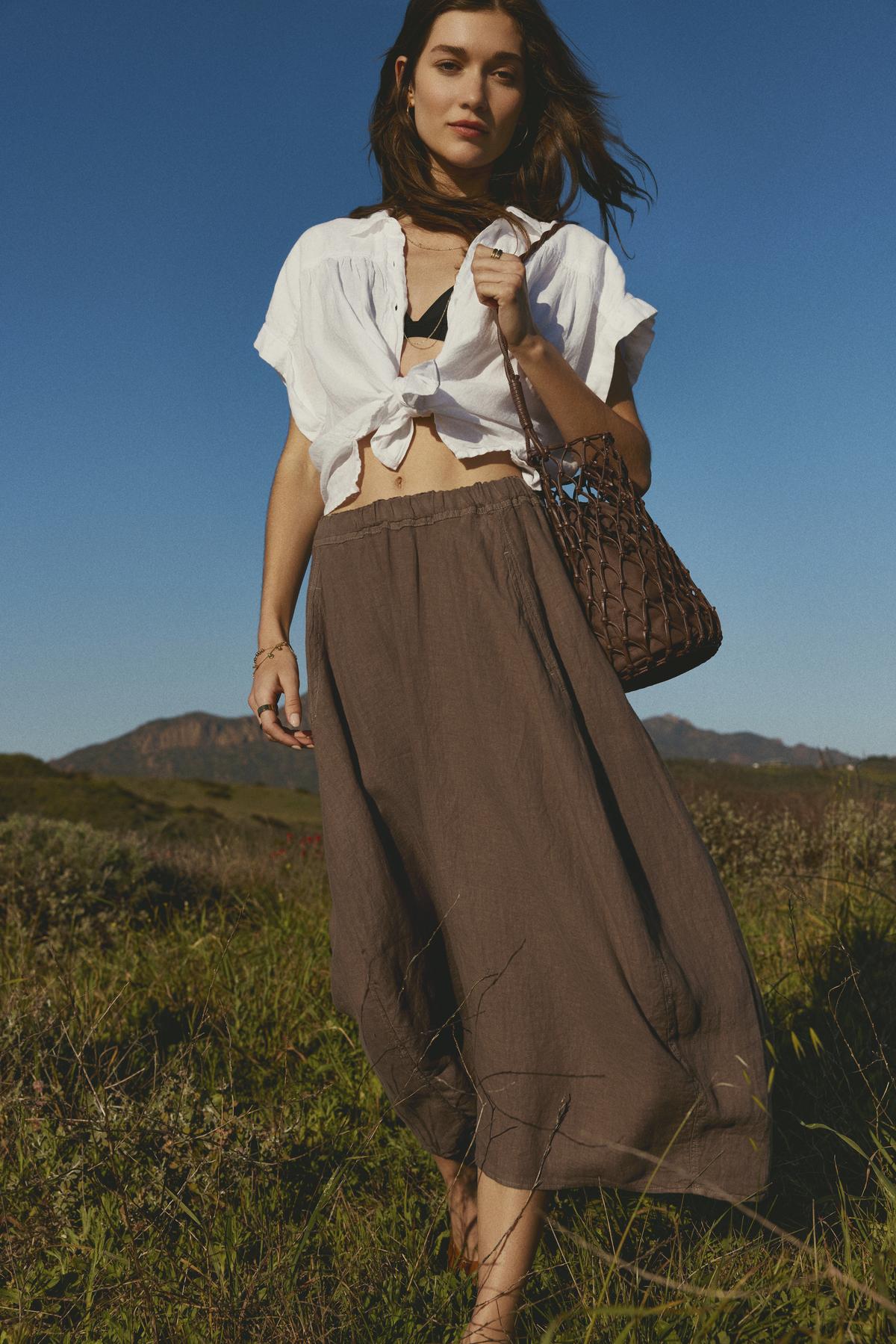   Woman walking in a field wearing a cropped ARIA LINEN BUTTON FRONT TOP from Velvet by Graham & Spencer, long brown skirt, and carrying a netted bag. 