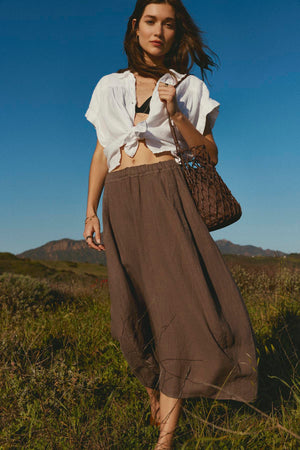 Woman walking in a field wearing a cropped ARIA LINEN BUTTON FRONT TOP from Velvet by Graham & Spencer, long brown skirt, and carrying a netted bag.