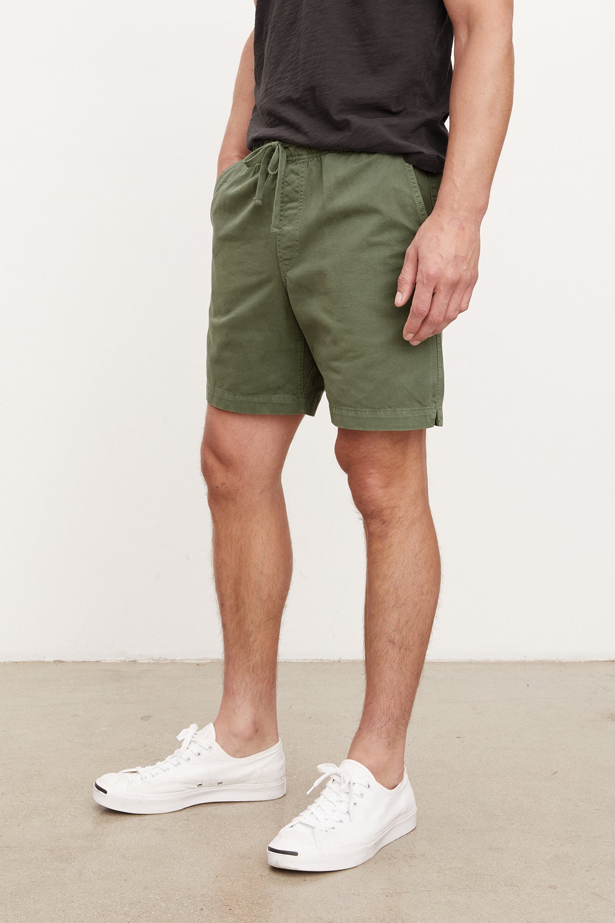   Man standing in a neutral pose wearing Velvet by Graham & Spencer's FIELDER SHORT, an olive green cotton twill shorts with an elastic drawstring waist, a black t-shirt, and white sneakers against a plain background. 