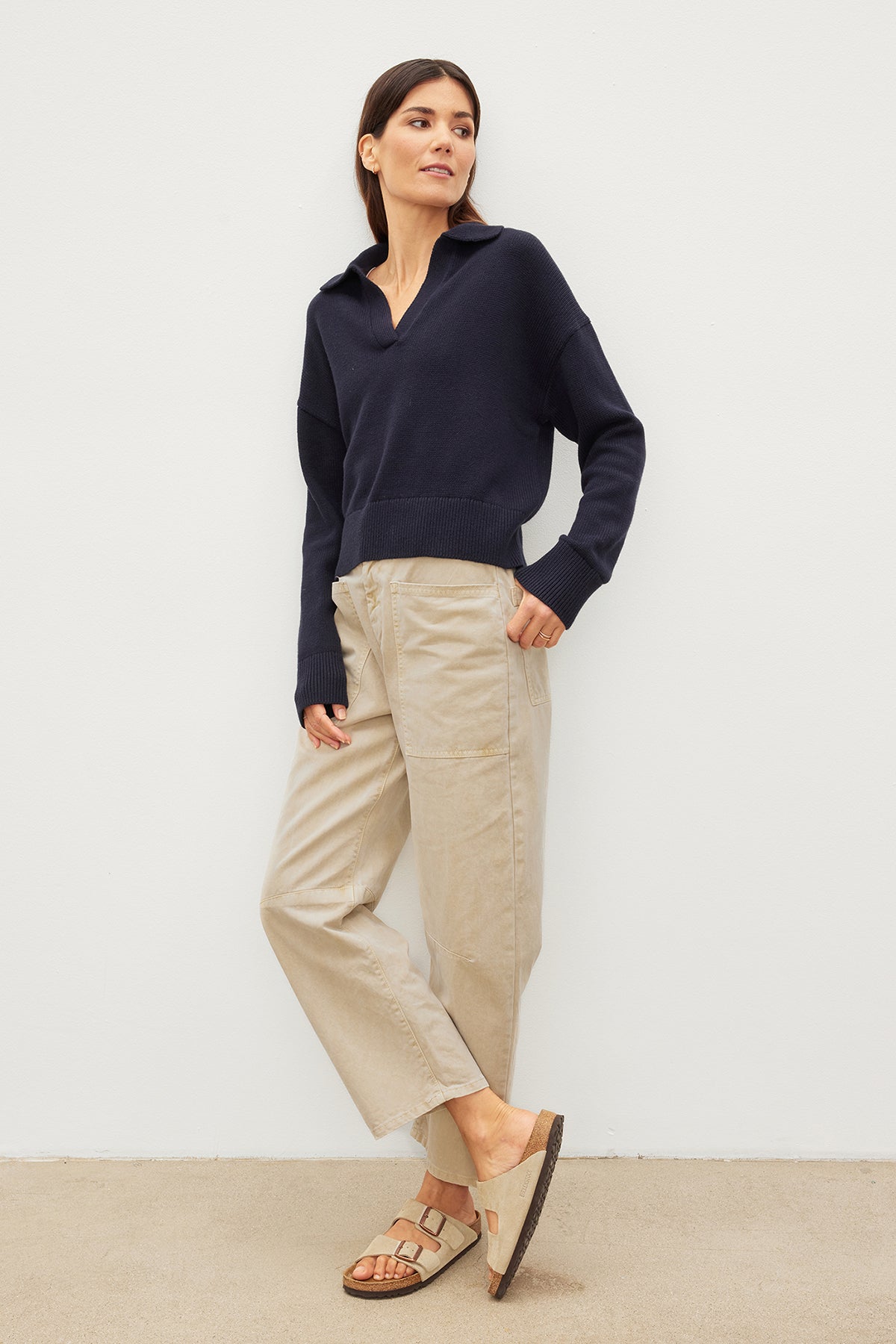   Woman in a navy blue sweater and beige Velvet by Graham & Spencer BRYLIE SANDED TWILL UTILITY PANT leaning against a white wall. 