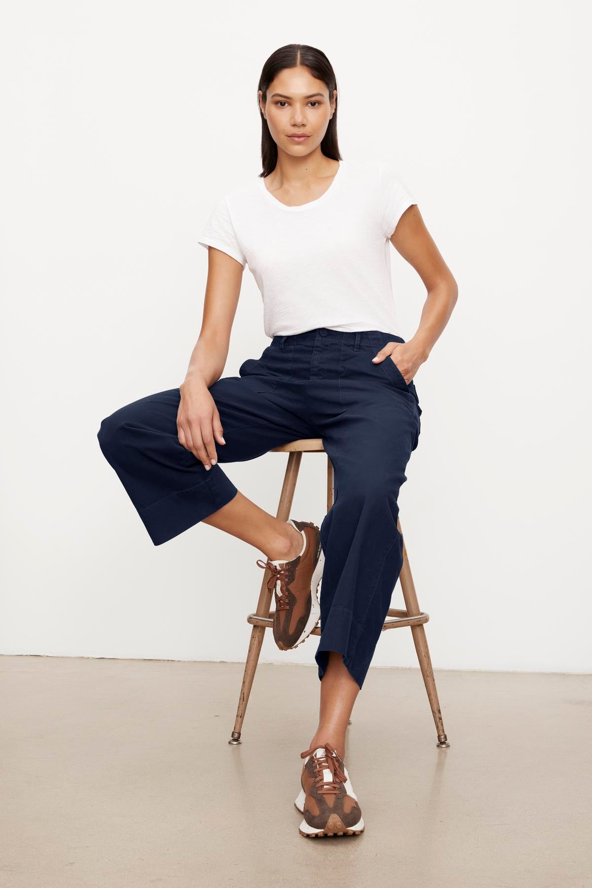   A woman sits on a wooden stool, wearing a white T-shirt and navy blue garment-dyed Velvet by Graham & Spencer culottes with brown sandals, facing the camera in a studio setting. 