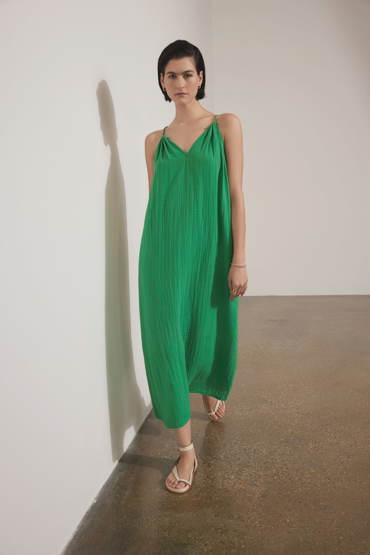 A woman wearing a flowing green Carrillo dress by Velvet by Jenny Graham and sandals stands against a white wall, casting a long shadow.-36863272026305