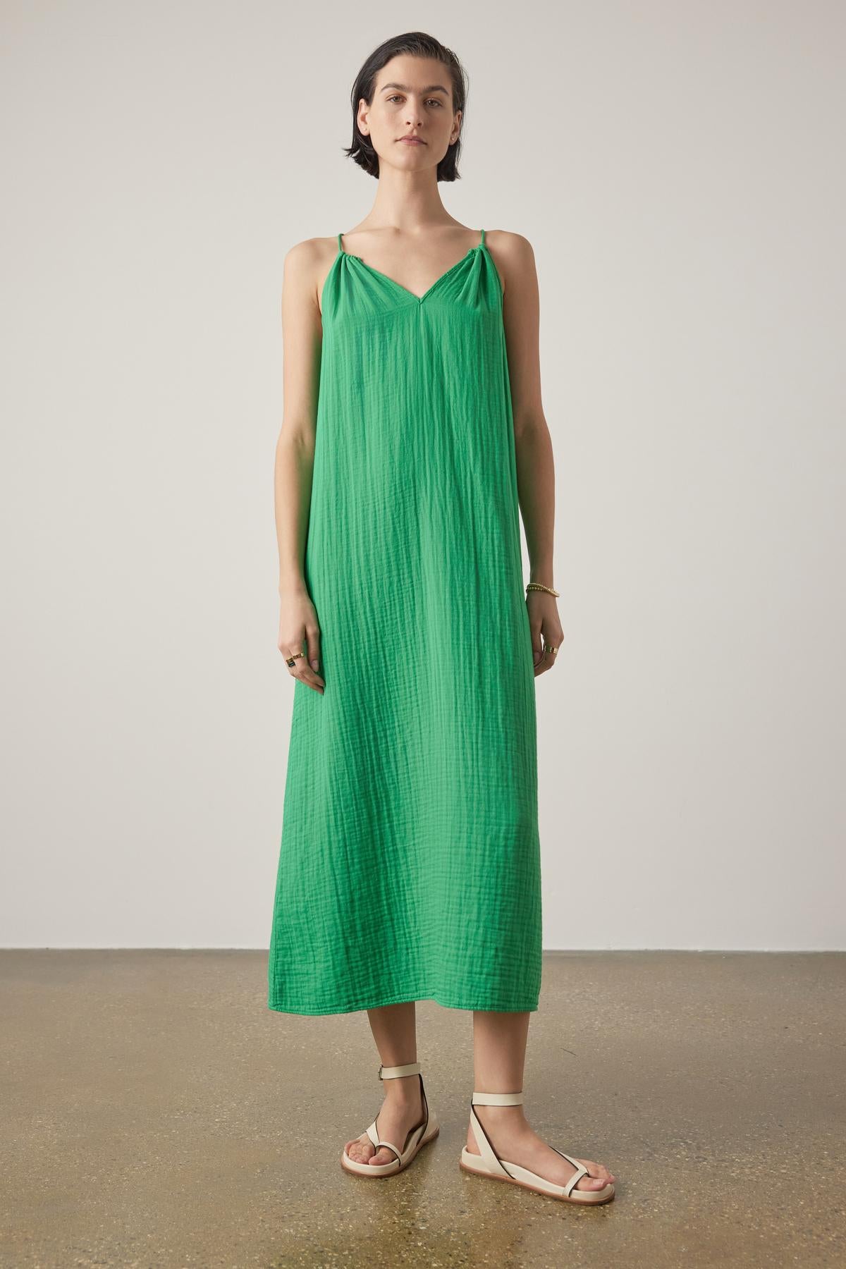 A woman stands wearing a CARRILLO DRESS by Velvet by Jenny Graham, featuring a bright green, pleated maxi design with thin straps. She has paired this dress with light-colored flat sandals against a neutral background.-36863272091841