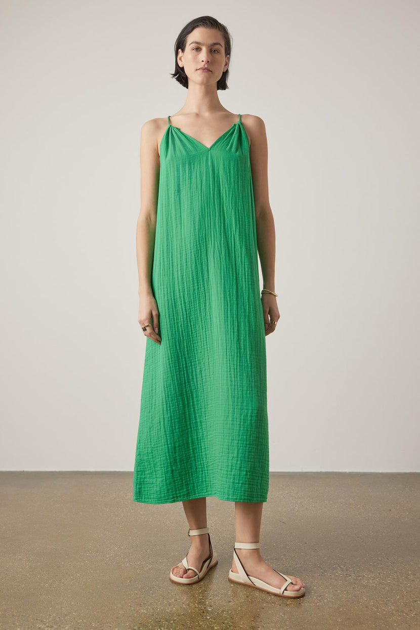 A woman stands wearing a CARRILLO DRESS by Velvet by Jenny Graham, featuring a bright green, pleated maxi design with thin straps. She has paired this dress with light-colored flat sandals against a neutral background.