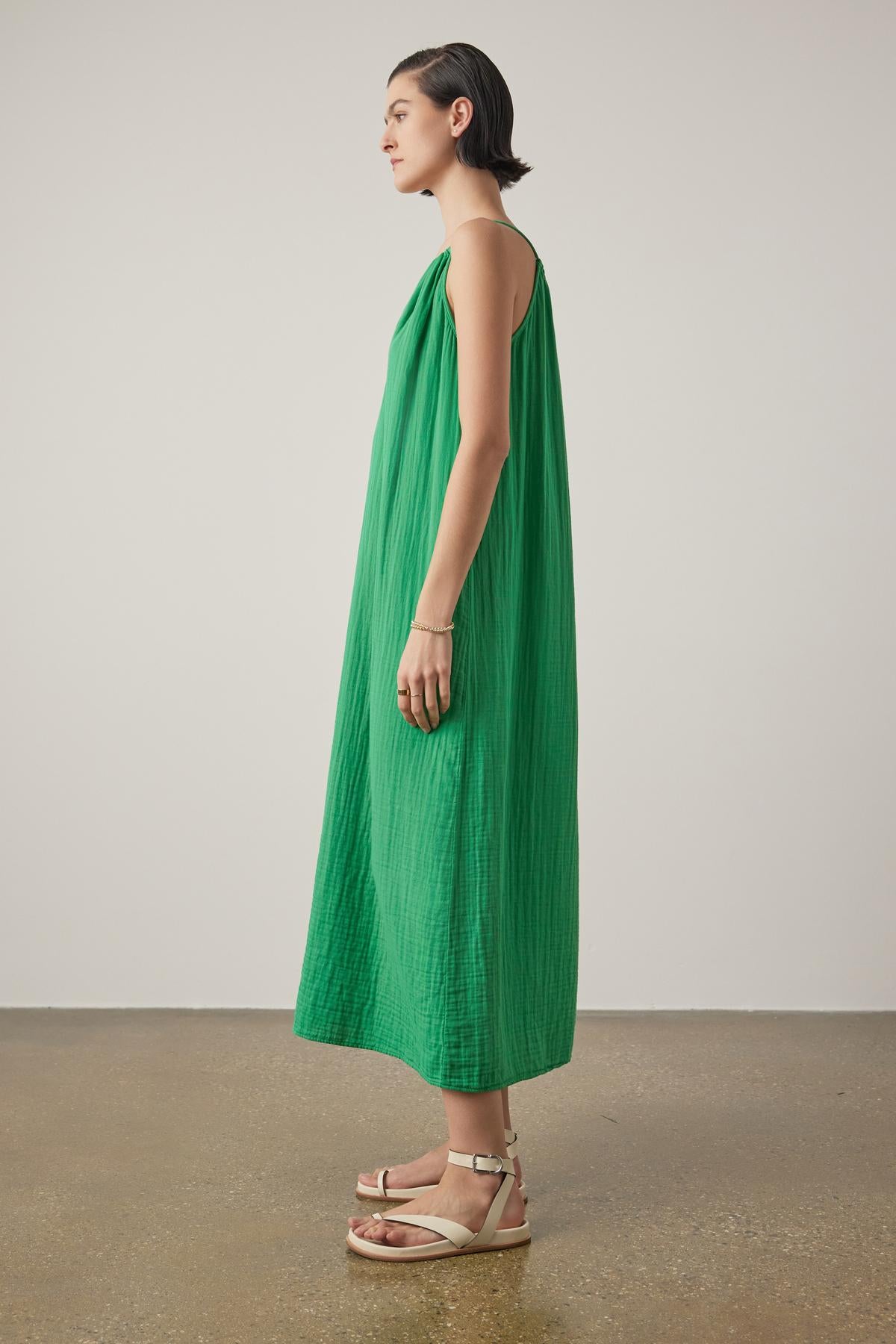 A woman standing in profile, wearing a long green sleeveless Carrillo dress by Velvet by Jenny Graham and beige sandals, against a neutral background.-36863272124609
