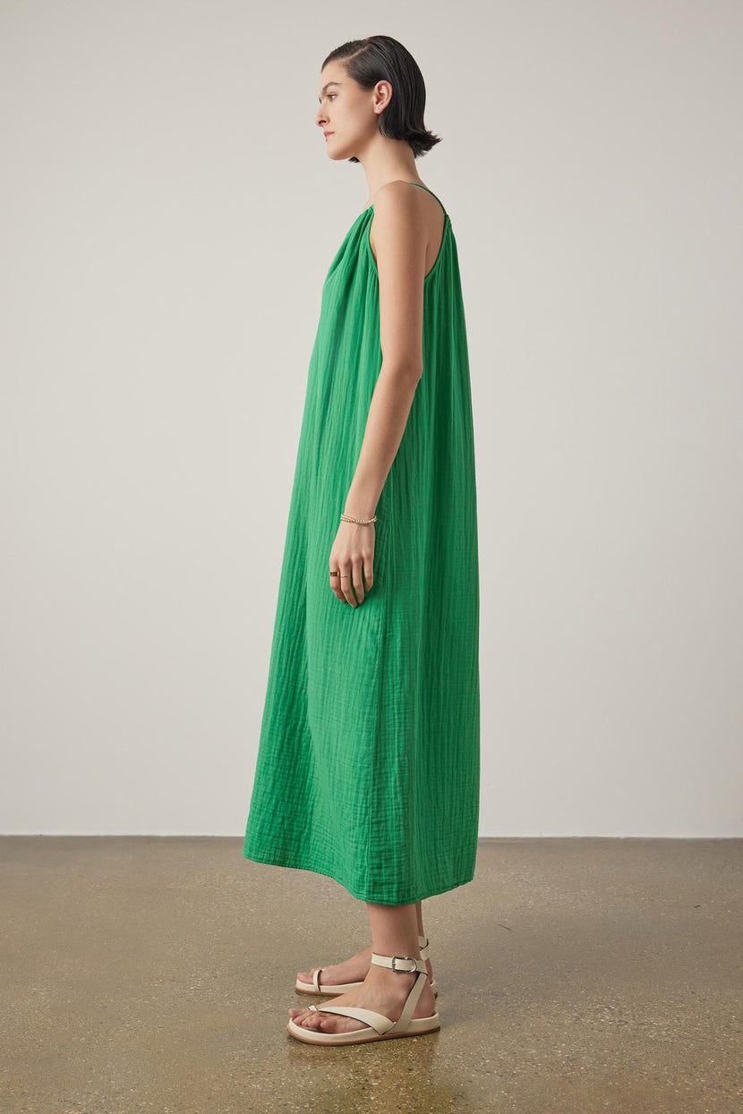 A woman standing in profile, wearing a long green sleeveless Carrillo dress by Velvet by Jenny Graham and beige sandals, against a neutral background.