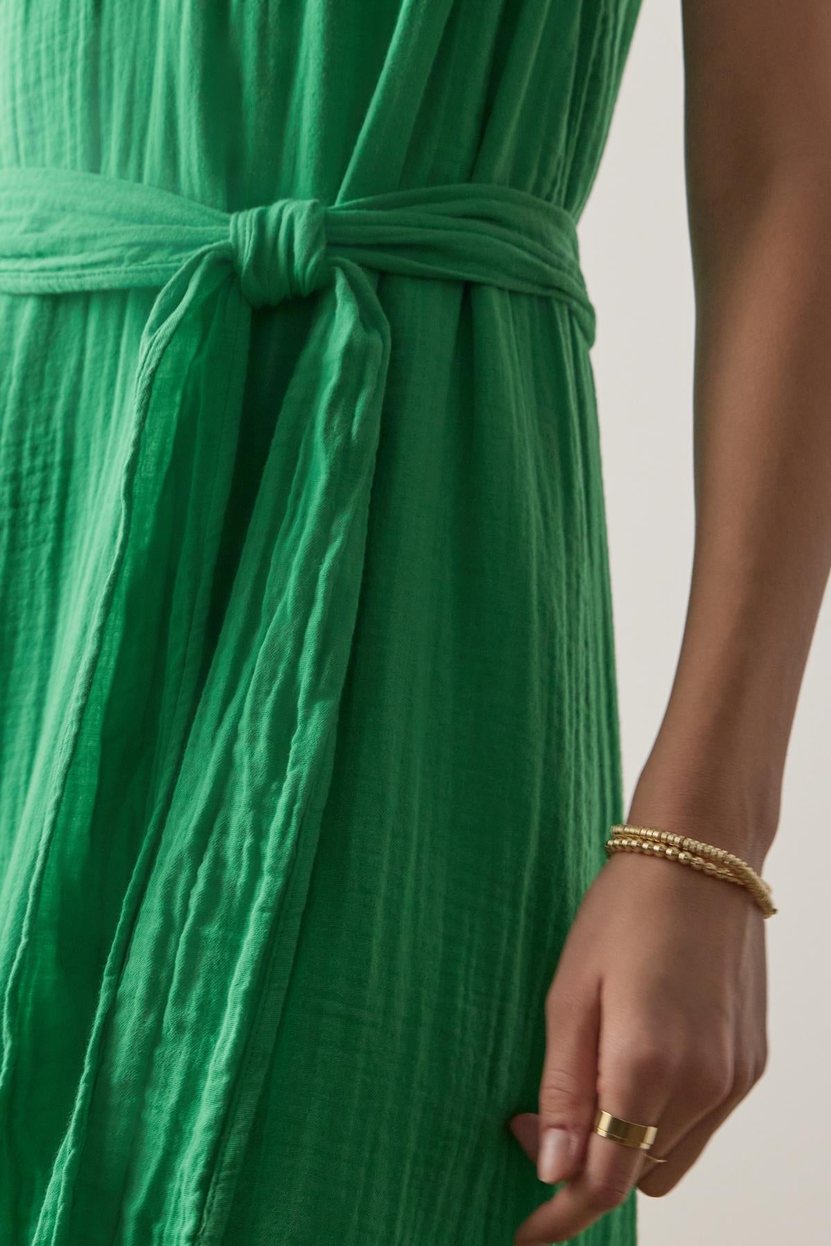 Close-up view of a person wearing a green cotton gauze Carrillo dress by Velvet by Jenny Graham with a knotted waist detail, accessorized with gold bracelets.-36863272059073