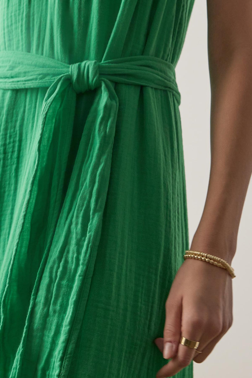 Close-up view of a person wearing a green cotton gauze Carrillo dress by Velvet by Jenny Graham with a knotted waist detail, accessorized with gold bracelets.