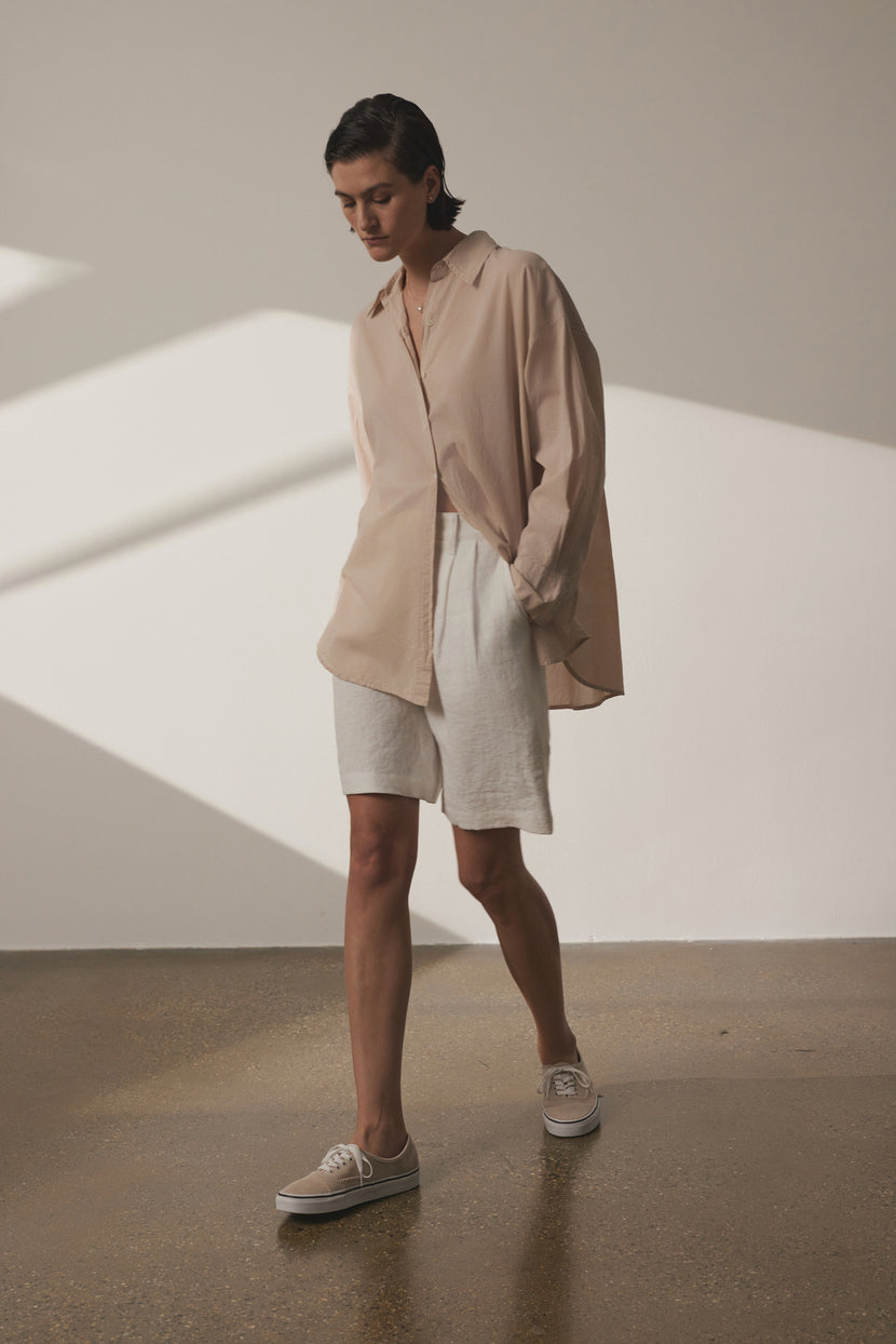 A person stands indoors, wearing an oversized Velvet by Jenny Graham REDONDO button-up shirt, white shorts, and tan sneakers, looking down thoughtfully in a sunlit room with shadows on the wall.