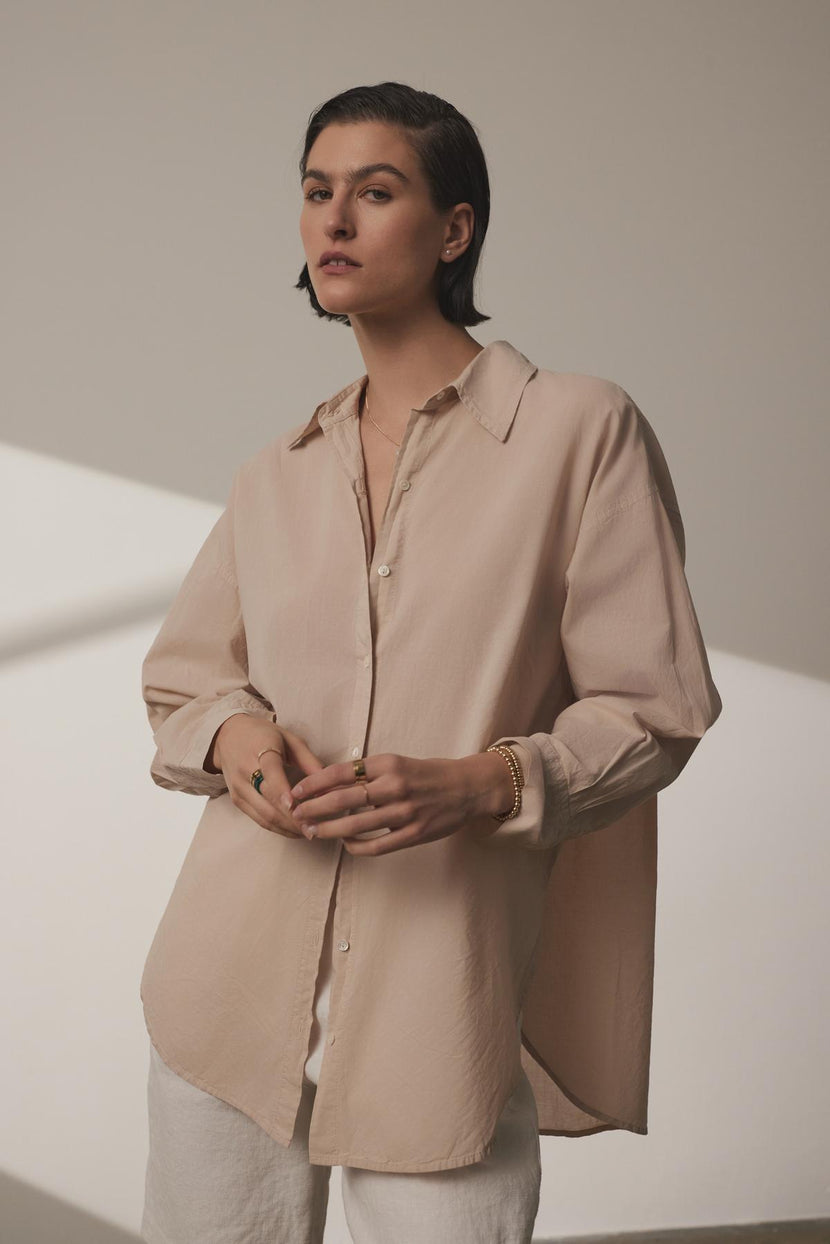 A woman in a Velvet by Jenny Graham REDONDO BUTTON-UP SHIRT and white relaxed trousers, standing with her hands clasped in front of her, against a softly lit beige background.