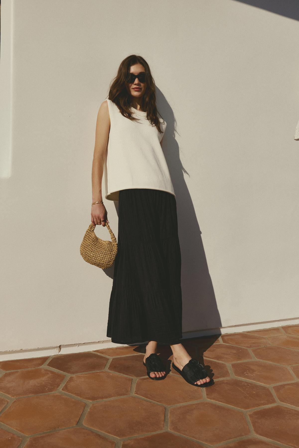   Woman in a ASTER CREW NECK SWEATER by Velvet by Graham & Spencer with ribbed detailing and black skirt standing against a white wall with a wicker bag. 