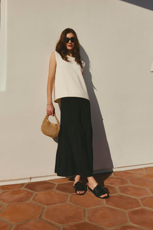 Woman in a ASTER CREW NECK SWEATER by Velvet by Graham & Spencer with ribbed detailing and black skirt standing against a white wall with a wicker bag.