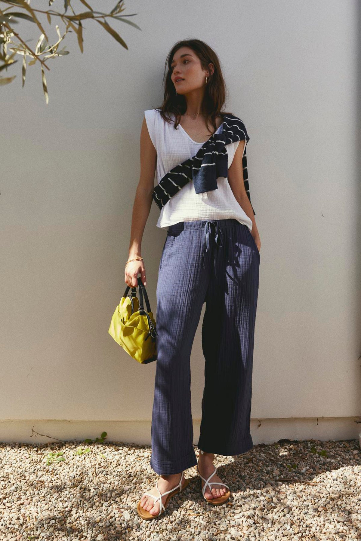   A woman in a JAYLA COTTON GAUZE TANK TOP by Velvet by Graham & Spencer and blue striped pants stands against a light wall, holding a yellow bag, with sunlight casting shadows of leaves. 