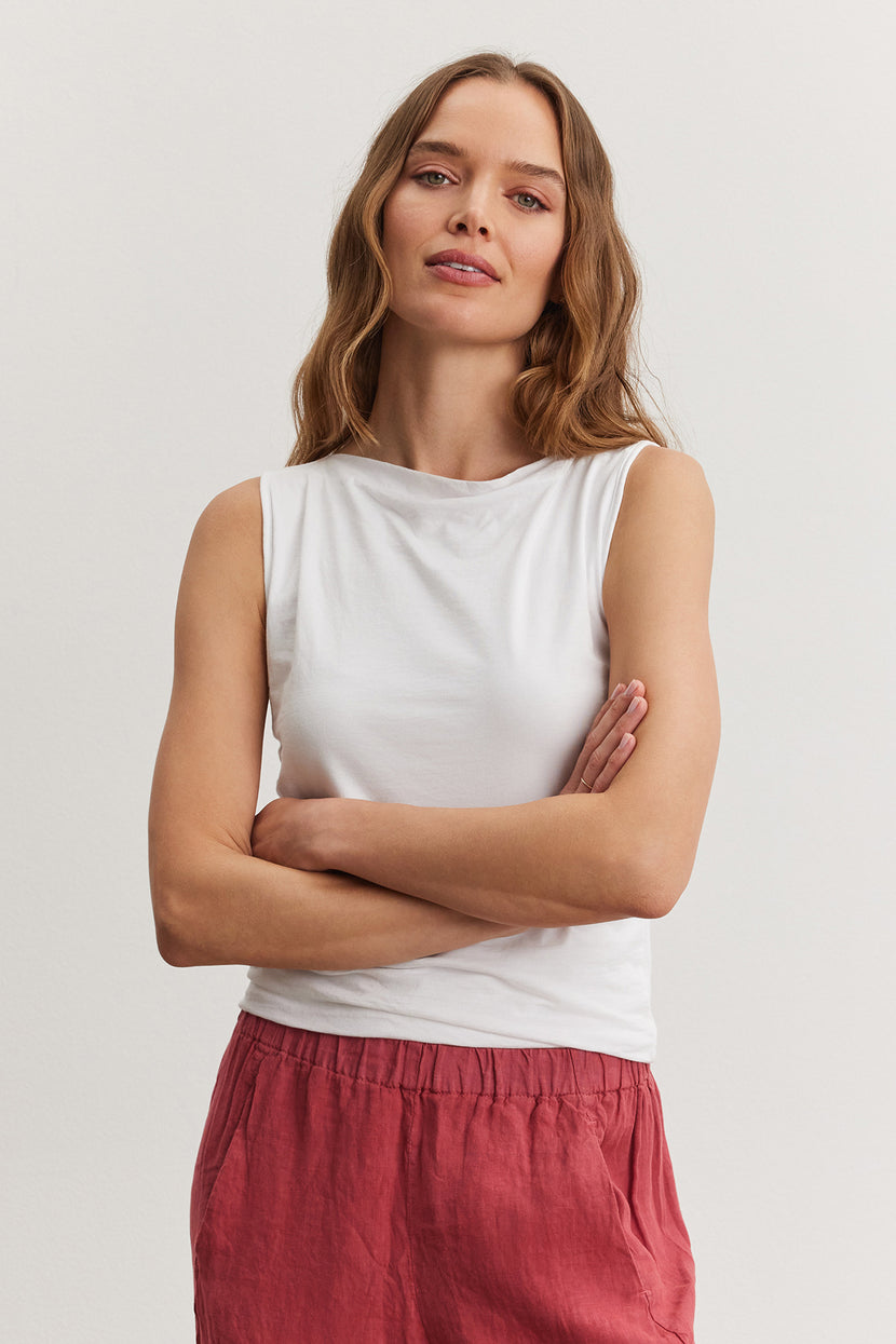 A woman with medium-length hair, wearing a Velvet by Graham & Spencer EMILIA TANK TOP and red pants, stands with her arms crossed.