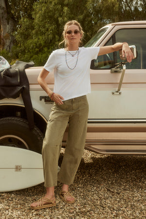 Woman in sunglasses, white t-shirt, and Velvet by Graham & Spencer BRYLIE SANDED TWILL UTILITY PANT leaning on a vintage pickup truck in a gravel area.