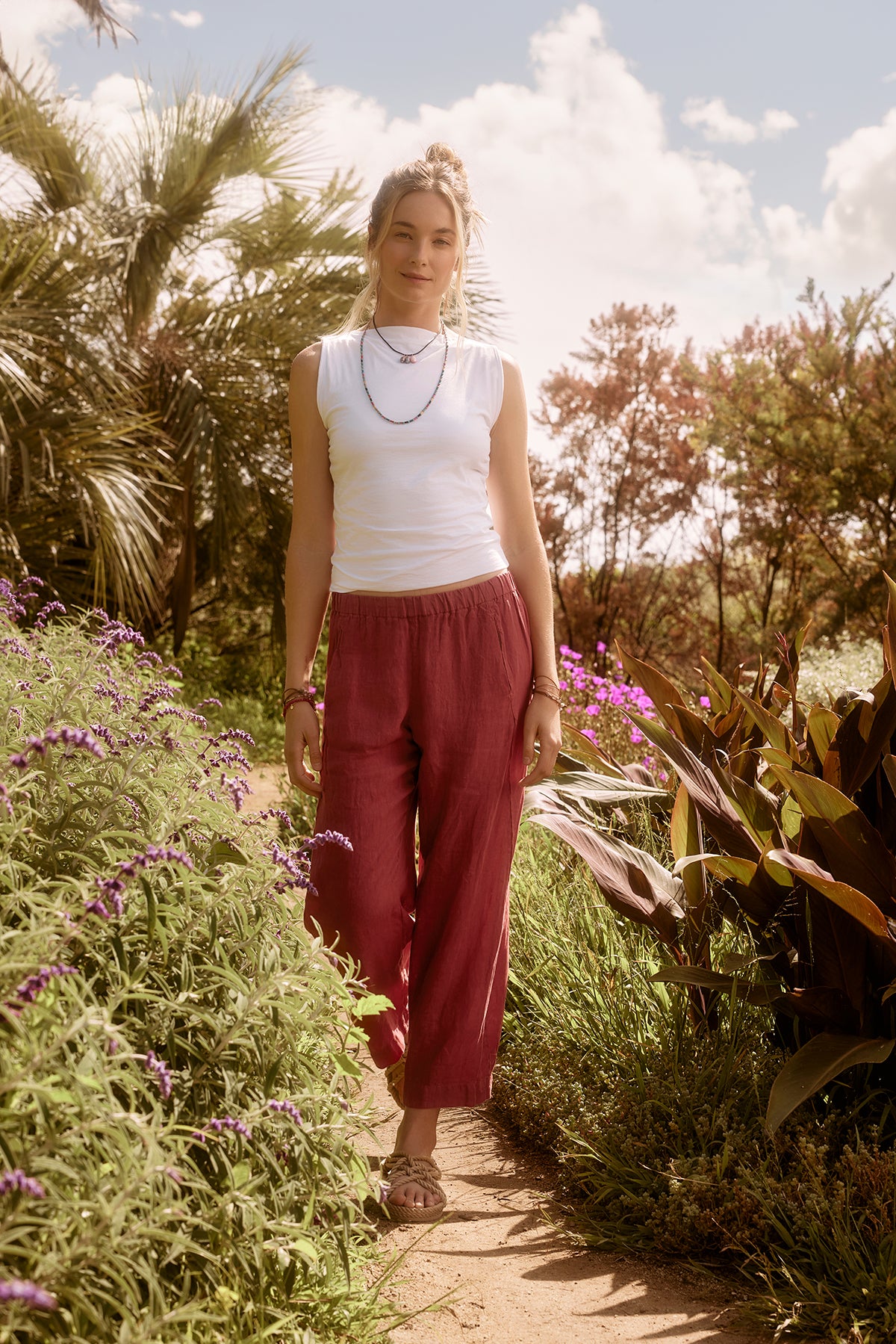 Woman standing on a garden path surrounded by purple flowers, wearing Velvet by Graham & Spencer's LOLA LINEN PANT and a white tank top, with sunlight filtering through trees.-36909780271297