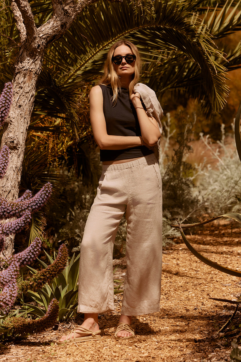 A woman in sunglasses stands under a tree in a garden, wearing beige trousers and an EMILIA TANK TOP by Velvet by Graham & Spencer, with her arms crossed.