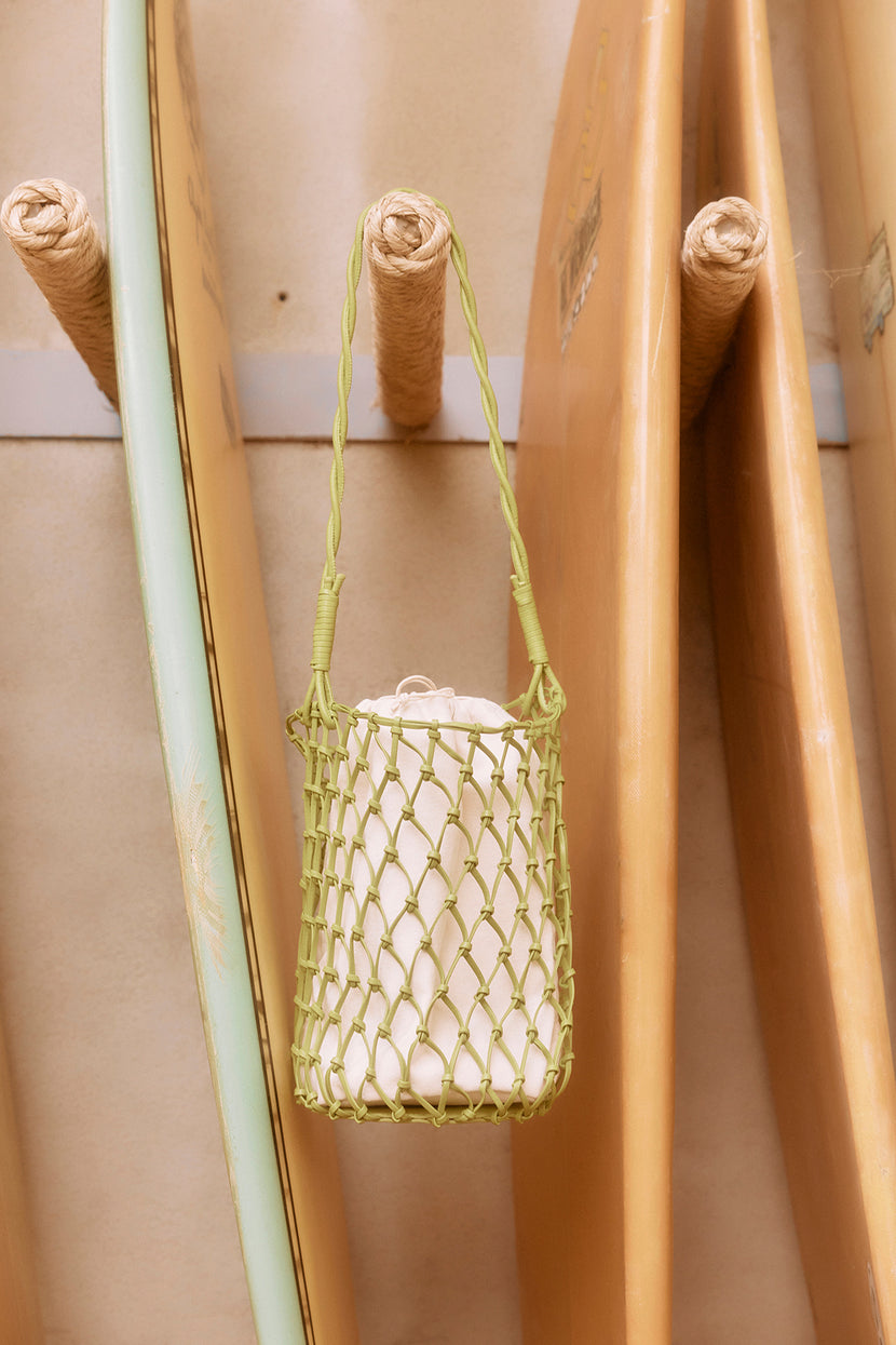 A green Velvet by Graham & Spencer mesh tote bag hanging from a wooden bamboo pole, with other bamboo poles in the background.