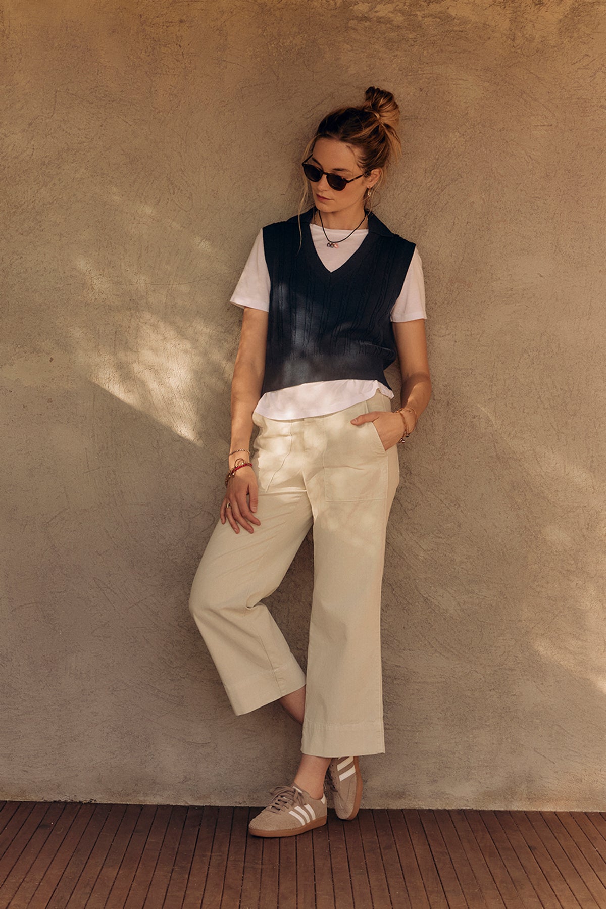   A stylish woman in sunglasses, a black vest, white shirt, and Velvet by Graham & Spencer MYA COTTON CANVAS PANT leans against a beige wall, hands in pockets. 