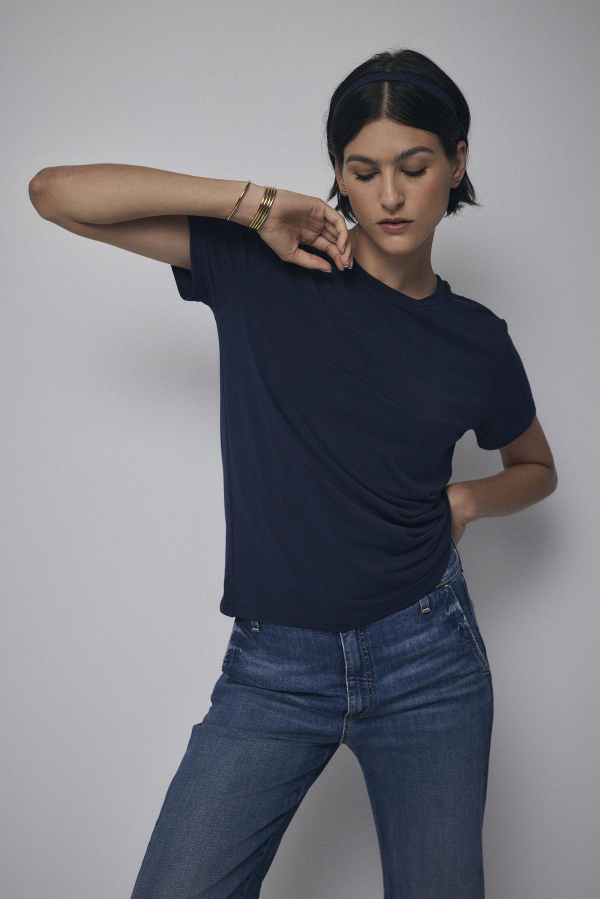   A woman in a navy blue Velvet by Jenny Graham Solana Tee and jeans poses with her hand on her waist, looking downward. 
