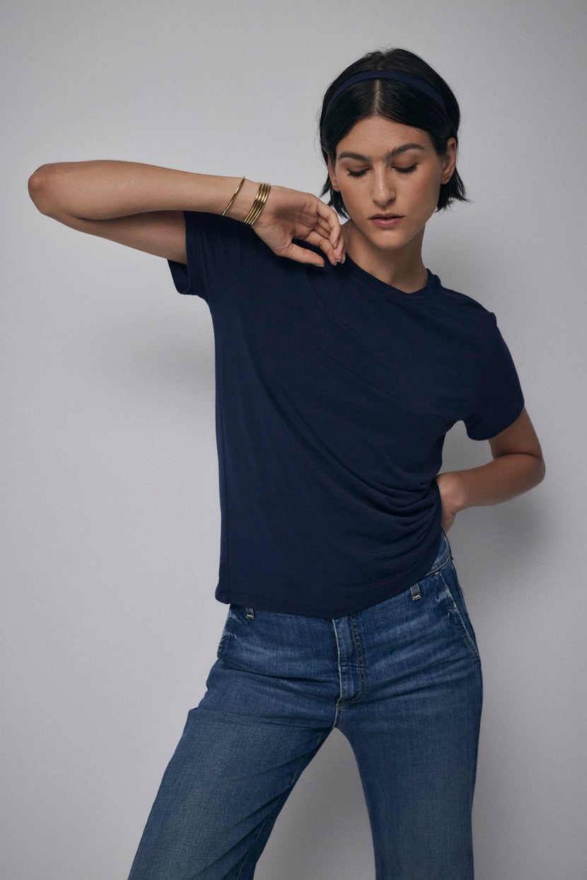 A woman in a navy blue Velvet by Jenny Graham Solana Tee and jeans poses with her hand on her waist, looking downward.