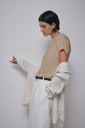 A person wearing a neutral-toned outfit with a Velvet by Jenny Graham SOLANA TEE draped over their arms, standing against a plain background and looking down at their hand.