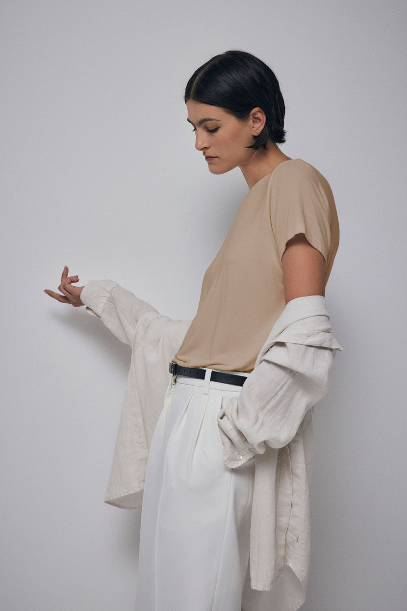 A person wearing a neutral-toned outfit with a Velvet by Jenny Graham SOLANA TEE draped over their arms, standing against a plain background and looking down at their hand.