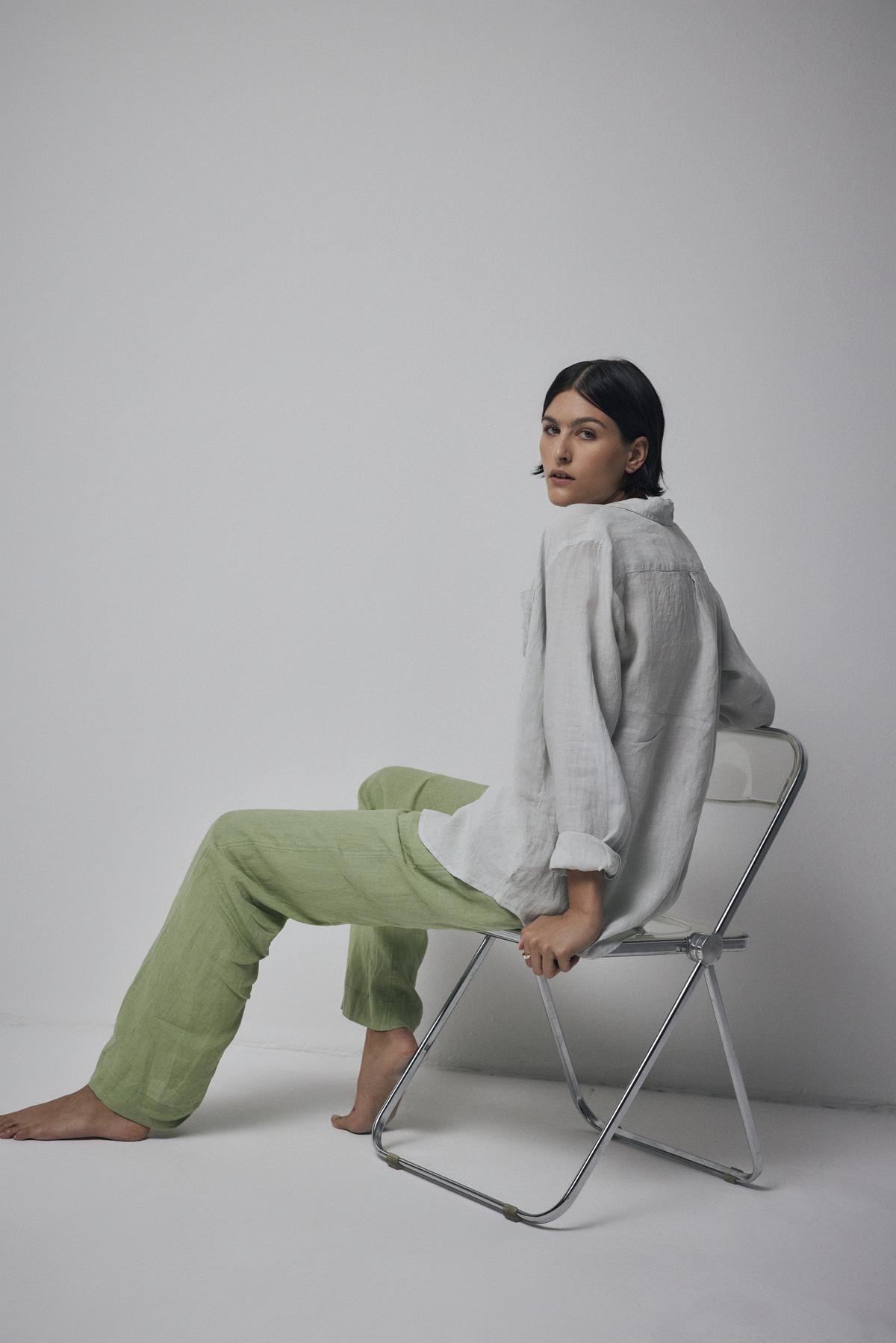   A person with long dark hair, dressed in a relaxed silhouette featuring a light-colored MULHOLLAND LINEN SHIRT by Velvet by Jenny Graham and green pants, sits barefoot on a folding chair against a plain backdrop. 