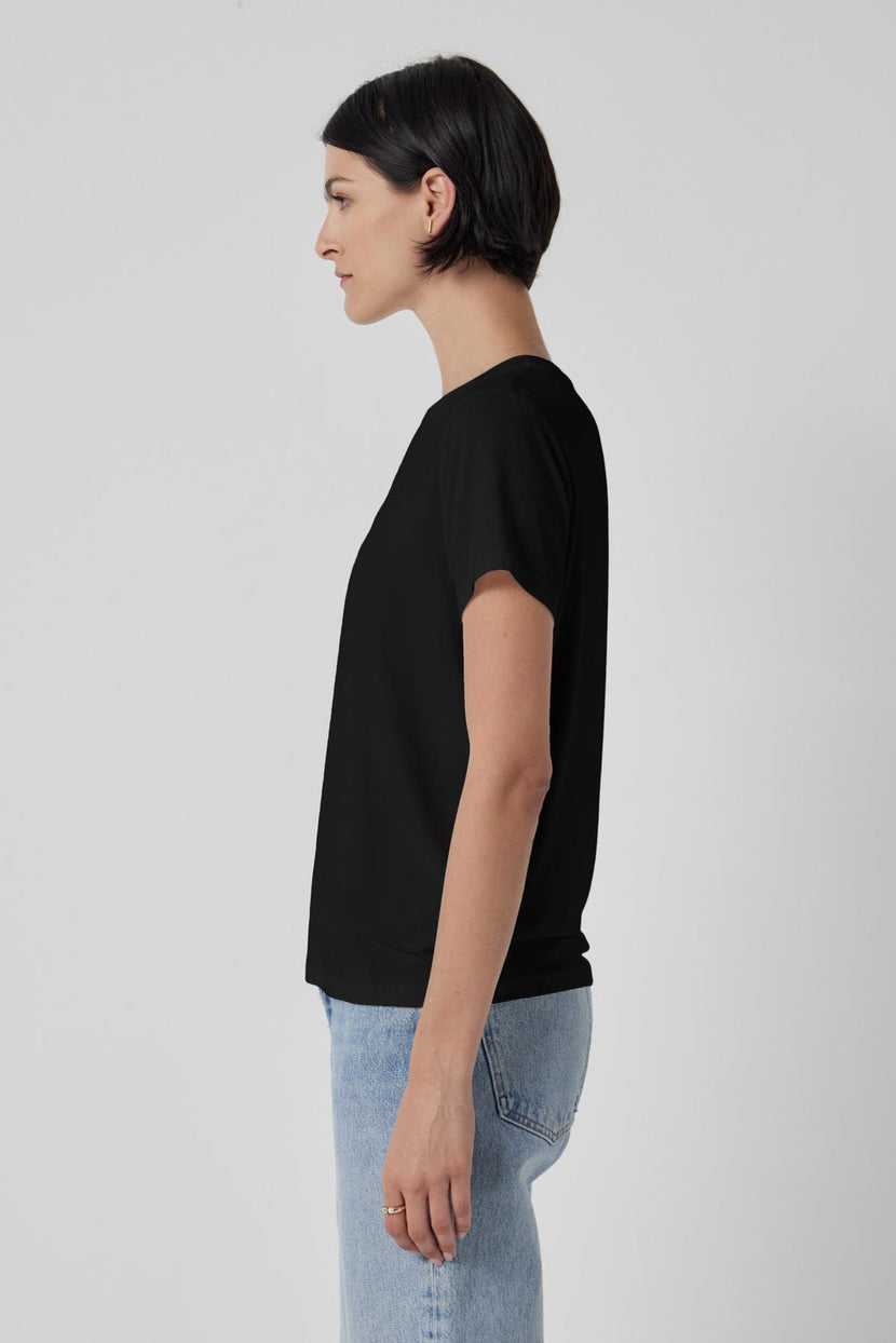 Woman standing in profile wearing a classic Solana Tee by Velvet by Jenny Graham and blue jeans against a gray background.