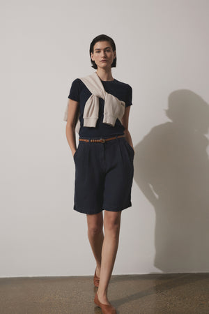 A woman in a dark blue skirt and a Velvet by Jenny Graham BEDFORD TEE stands confidently in a studio, with her shadow visible on the wall.