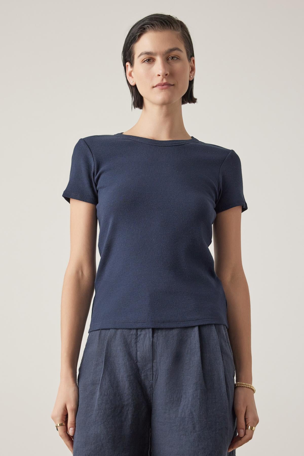 A woman with short hair, wearing a Velvet by Jenny Graham dark blue Bedford Tee with a crew neckline and gray pants, stands against a light background.-36753616863425