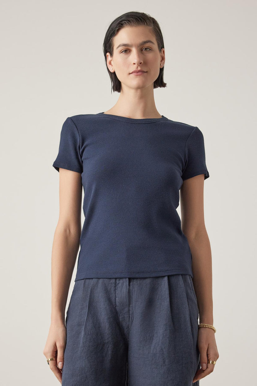 A woman with short hair, wearing a Velvet by Jenny Graham dark blue Bedford Tee with a crew neckline and gray pants, stands against a light background.