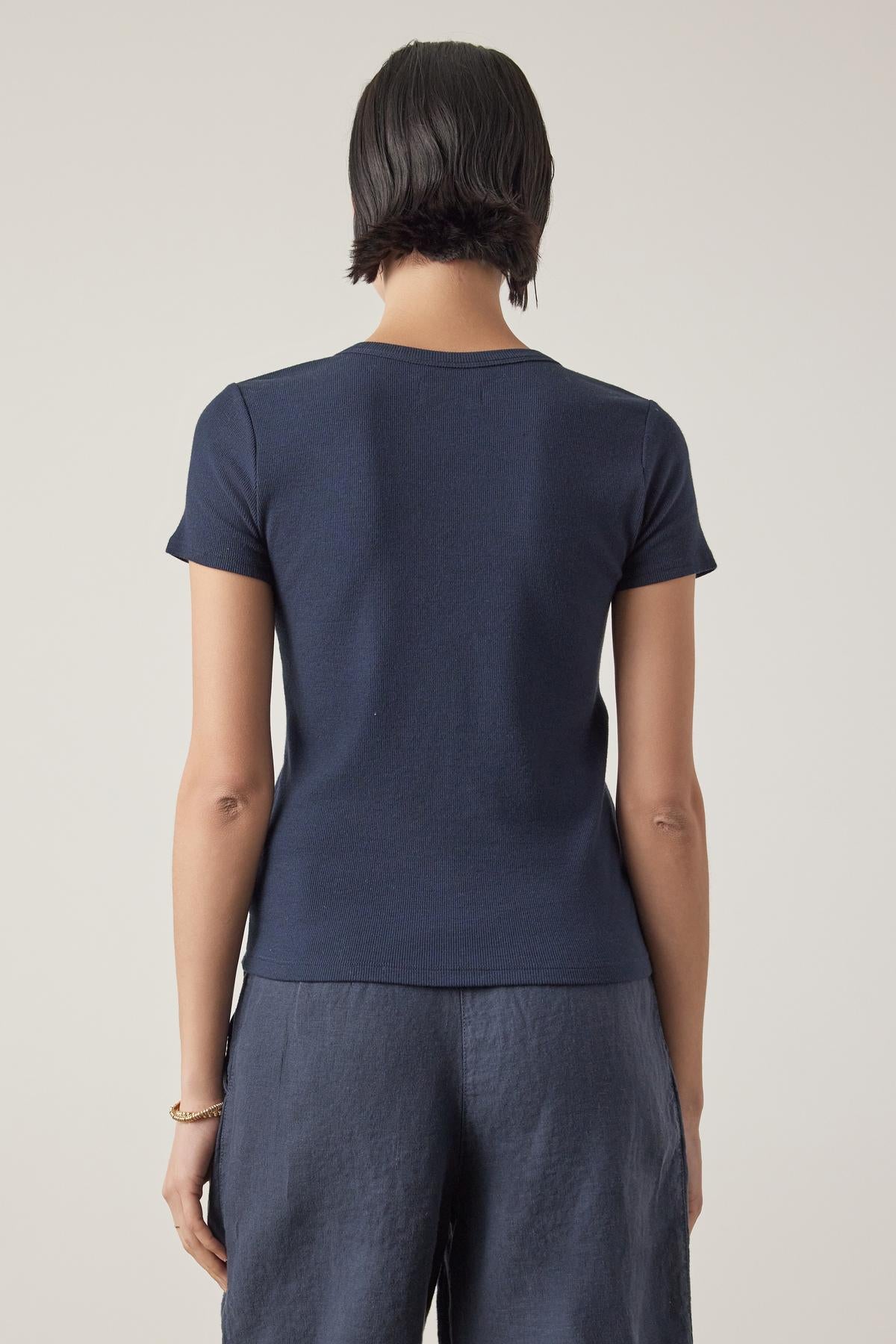 Rear view of a woman in a Velvet by Jenny Graham BEDFORD TEE, a dark blue short-sleeved top with a crew neckline, and grey pants, standing against a light beige background.-36753616928961