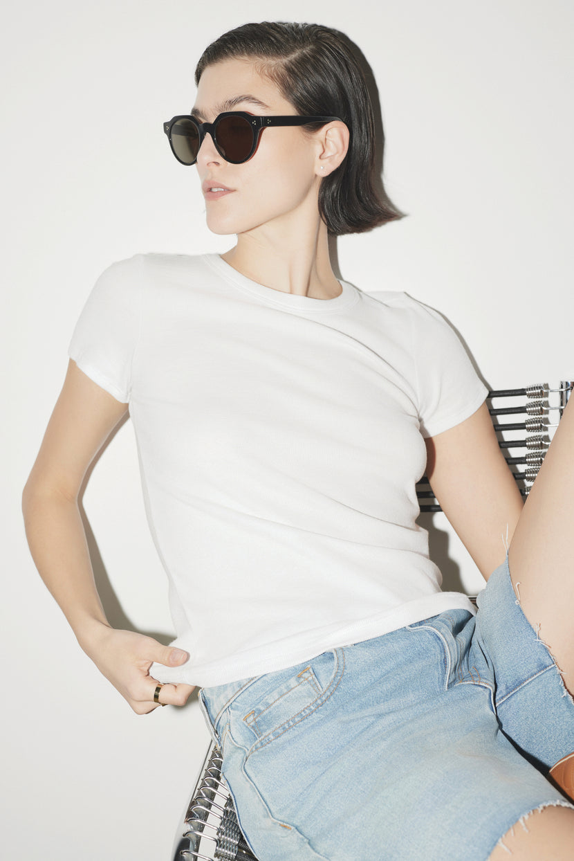 A woman in sunglasses, a white Velvet by Jenny Graham BEDFORD TEE with a crew neckline, and jeans, sitting on a metal chair against a white background, looking to the side.